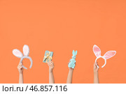 Женские руки держат ободки с ушками Пасхального кролика с подарочной коробкой и игрушечным кроликом на оранжевом фоне. Стоковое фото, фотограф Pixel_shot / Фотобанк Лори