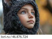 A girl with blue eyes and fair skin covered with freckles. She is wearing a soft, fluffy headdress with earflaps, reminiscent of a hood in the form of a woolen animal costume, perhaps a cat or a wolf. Her gaze is directed forward, creating a sense of dreaminess and thoughtfulness. The background is blurred and contains warm autumnal tones, which adds depth and softness to the image. The lighting is natural and emphasizes the texture of the fur and the delicacy of the child's facial features. Стоковое AI-изображение, AI-художник Анна Кривицкая / Фотобанк Лори