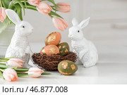 A bouquet of delicate tulips, Easter bunnies and eggs with a golden pattern on the table. There is a white kitchen in the background. Beautiful greeting card. The concept of minimalism. Стоковое фото, фотограф Photojuli / Фотобанк Лори