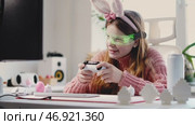Easter meets gaming as young girl plays with enthusiasm, holding controller with excitement. Decorative eggs on desk add seasonal charm, blending traditional holiday spirit with modern gaming culture. Стоковое видео, видеограф Енваев Андрей / Фотобанк Лори