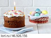 Традиционный пасхальный кулич, празднично украшенный глазурью и разноцветной сладкой обсыпкой, с горящей свечой. Крашеные яйца в плетеной корзине на заднем плане в расфокусе. Стоковое фото, фотограф Светлана Ельцова / Фотобанк Лори