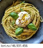 A plate of pasta topped with a fragrant pesto of basil, nuts and cheese. In the center of the dish lies a delicious ball of fresh burrata, drizzled with olive oil and sprinkled with ground black pepper. Fresh basil leaves decorate the dish, adding a bright green accent. A sprinkle of crushed nuts and cheese adds textural contrast to the delicate pasta.The color palette of the photo highlights the natural tones of the ingredients - the golden color of the spaghetti, the green of the pesto, the creamy shade of the burrata and the dark background of the plate create a stylish and appetizing look. Стоковое AI-изображение, AI-художник Анна Кривицкая / Фотобанк Лори