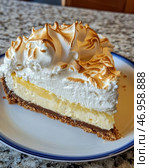 A delicious slice of Lemon Meringue Pie on a plate. The dessert consists of several layers: a crisp shortbread base, delicate lemon curd, and airy meringue with golden charred marks created by caramelization. The top layer of meringue looks fluffy and light, with beautiful waves adding texture. The pie is served on a white plate with a blue border, and a granite countertop is visible in the background. The atmosphere of the image conveys a sense of coziness and pleasure from a sweet treat. Стоковое AI-изображение, AI-художник Анна Кривицкая / Фотобанк Лори