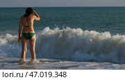Rear view of a young beautiful girl in bikini standing on sea shore and watching the coming waves. Стоковое фото, фотограф David Kashakhi / Фотобанк Лори