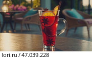 A tall transparent glass mug filled with deep red hibiscus iced tea, garnished with orange slices, star anise, and a cinnamon stick, placed on a table with a blurred exotic patio in the background. Стоковое AI-видео, аниматор Башкирова Анна / Фотобанк Лори