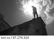 Young man standing on the edge of an abandoned building, silhouetted against a bright sun surrounded by clouds. The shot is taken from below, enhancing the sense of height and the contrast between the man and the sky. The atmosphere is tense, ambiguous, perhaps symbolizing a search for meaning, risk, or overcoming fears. The buildings in the background emphasize the urban nature of the scene, adding a sense of loneliness or contemplation of the future. Стоковое AI-изображение, AI-художник Анна Кривицкая / Фотобанк Лори