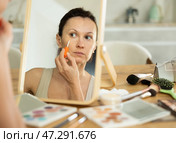 Middle-aged woman applying concealer on her facial skin sitting in front of the mirror. Стоковое фото, фотограф Яков Филимонов / Фотобанк Лори