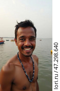 A cheerful man stands near the Ganges River in Varanasi, showcasing a genuine smile. The backdrop features boats floating on tranquil water, reflecting the beauty of daily life Generative AI. Стоковое AI-изображение, AI-художник Юлия Петровская / Фотобанк Лори