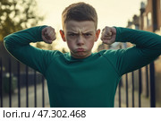 Kid boy shows his muscles. Angry little child boy waves his fist at the camera outdoors at the city street. Unhappy grumpy boy showing his biceps. Provocation. Elementary school problems. Bullying. Стоковое AI-изображение, AI-художник Марина Демидюк / Фотобанк Лори