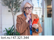 Thoughtful senior woman with smartphone looking away in an urban environment. Стоковое фото, фотограф Raquel Corcoles Mendez / Фотобанк Лори