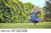 Young blonde girl spinning playfully in white sundress, dancing barefoot on lush green lawn near garden hedgerow, radiating childhood joy. Стоковое фото, фотограф Mikhail Davidovich / Фотобанк Лори