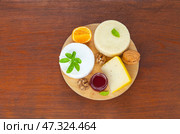 Three kinds of cream cheeses with basil, mandarin, fruit jam and walnuts on round cutting board on wooden table. Organic nutritious food. Top view, close-up, flat lay, copy space, mock up. Стоковое фото, фотограф Виктория Катьянова / Фотобанк Лори