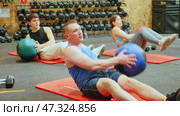 Focused adult man taking part in high-intensity group training session, doing V-sit exercise with medicine ball and twisting torso to strengthen abdominal and oblique muscles. Стоковое видео, видеограф Яков Филимонов / Фотобанк Лори