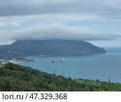 A picturesque view of Gurzuf from above. In the center of the composition is the majestic Mount Ayu-Dag, covered with a thick cap of clouds, like a magic veil. Below is the coast with houses, hotels and a bay in which the famous Adalary rocks are visible, like two guards at the entrance to the sea. Everything is surrounded by dense Crimean forests and greenery, creating a feeling of silence, peace and harmony. The atmosphere is filled with cool freshness, the breath of the sea and the anticipation of something new. Стоковое фото, фотограф Анна Кривицкая / Фотобанк Лори