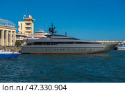 A fragment of the seaport in Yalta. In the center of the frame, an elegant and modern yacht called Orion is moored, painted dark gray with an orange line along the waterline. Nearby are boats and other vessels, and in the background are visible buildings of the port infrastructure, including the control tower with a characteristic dome. The sea is bright and rich blue, light ripples on the water indicate a fresh breeze. On the concrete wall behind is the inscription WELCOME, which emphasizes the tourist character of the place.The photo conveys the atmosphere of active marine life and the modern Yalta coast. (2021 год). Редакционное фото, фотограф Анна Кривицкая / Фотобанк Лори