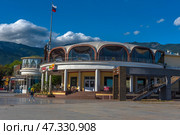 A fragment of the Yalta embankment, one of the most popular resort towns in Crimea. In the foreground is a modern building with a round façade and stained-glass windows, where cafes and restaurants are located. On the left is part of the pedestrian zone with passers-by, vacationers and strollers along the shore. In the background, the green mountains of the Crimean ridge rise, and the sky is decorated with living clouds and a flying bird. The Russian flag on the mast emphasizes the formality and maritime character of the place.The decor conveys an atmosphere of relaxation, resort activity and harmony with the natural landscape. (2021 год). Редакционное фото, фотограф Анна Кривицкая / Фотобанк Лори