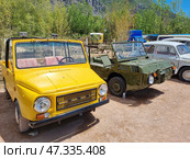 Two unusual open cars standing next to each other in an outdoor retro display.On the left is a bright yellow LuAZ-969 (possibly modified), a light Soviet open-top SUV with a steering wheel on the right, designed for off-road use. Its characteristic features are square shapes, simple optics, and high cross-country ability. The car looks unusual because of its paint and the textiles on the seats.On the right is a military green PT-S (floating transporter-snowmobile) or one of the early experimental amphibious vehicles of Soviet production. Its appearance is as utilitarian as possible: an open body, narrow tires, small seats, headlights placed outside, and a body slanted for movement on water. This car clearly had a military or special purpose.Other retro cars are visible behind, and the surroundings themselves are mountainous, possibly in Crimea. (2021 год). Редакционное фото, фотограф Анна Кривицкая / Фотобанк Лори