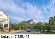 A picturesque corner of Crimea — a road running through green hills at the foot of a mountain, presumably the Kush-Kaya rock massif in the Simeiz region. A passenger car and a tractor with road equipment are moving along the highway. On the left, you can see a well-kept fence with forged elements, a building with an attic, and a billboard offering dishes from the cuisine. On the right and in the distance, there is dense vegetation, and a majestic rock rises above the entire landscape. The clear sky and soft light give the scene a warm, Crimean atmosphere. (2021 год). Редакционное фото, фотограф Анна Кривицкая / Фотобанк Лори