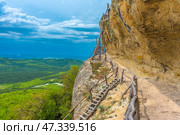One of the picturesque sections of the Chelter-Marmara cave monastery, located in the western part of Crimea, near Bakhchisarai. A narrow path with a wooden railing winds along a rocky cliff leading to a rough staircase made of unprocessed trunks and branches. The cliff seems to hang over the road, and beyond its edge is a bottomless valley and a majestic view of the Crimean foothills.This place is filled with the spirit of hermitage, peace and the grandeur of nature. A sign on the wall of the cliff warns of the height and calls for attention, reminding of the harsh but beautiful conditions in which the monks lived. Everything here - both the architecture and the rock landscape - is like a living testimony to time and the power of faith. Стоковое фото, фотограф Анна Кривицкая / Фотобанк Лори