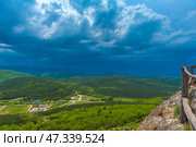 A breathtaking view from a mountain ledge in the Chelter-Marmara area of ​​Crimea. In the foreground is the edge of a cliff with a wooden fence, reminding us of the presence of a hiking trail and an observation deck. In the distance are endless green spaces covered with dense forests and smoothly turning into hills. The dramatic sky with heavy blue clouds creates a sense of the grandeur of nature, its power and mystery.This view seems to invite reflection, inspire and return to the inner silence that is so valuable in places like Chelter-Marmara - an ancient monastery hidden in the rocks. Стоковое фото, фотограф Анна Кривицкая / Фотобанк Лори