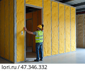 A construction worker in a safety vest examines the newly installed soundproofing material on the walls of an office space. The room is in progress. Стоковое AI-изображение, AI-художник Sergey Grishin / Фотобанк Лори