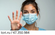 A young woman in a light blue outfit is wearing a face mask and raising her hand in a stop gesture. She appears to be in a medical or clinical environment during the pandemic. Стоковое AI-изображение, AI-художник Sergey Grishin / Фотобанк Лори