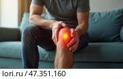 A man sits on a comfortable couch, holding his knee with a pained expression. The warm indoor lighting suggests it\'s daytime, indicating he might be dealing with knee pain or injury. Стоковое AI-изображение, AI-художник Sergey Grishin / Фотобанк Лори