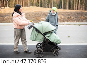 Женщина стоит с зеленой коляской, разговаривая со своим сыном-подростком. Семья в куртках идет по дороге весенним днем, мать, сын и коляска с младенцем. Стоковое фото, фотограф Яна Проничева / Фотобанк Лори