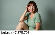 Joyful Woman Smiles Warmly While Seated in a Soft Green Outfit Against a Muted Backdrop, Embracing a Moment of Happiness and Self-Confidence. Стоковое AI-изображение, AI-художник Sergey Grishin / Фотобанк Лори