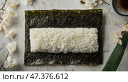Glossy nori sheet with sushi rice and floral accents. Глянцевый лист нори уложен ровным слоем, а сверху равномерно распределен тонкий, нежный слой риса для суши. Свежие цветочные акценты дополняют элегантную подачу, а рядом стоит чашка чая. Стоковое AI-изображение, AI-художник Katsiaryna Repich / Фотобанк Лори