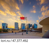 Doha, Qatar - Jan 06 2024, a view of flagpole with the flag of Qatar, fluttering in the wind, with the National Museum of Qatar on background, at evening with cloudy sky, Doha, Qatar. Редакционное фото, фотограф Сергей Богомяко / Фотобанк Лори