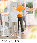 Dancing positive elderly women and men engaged in group class perform exercise near ballet barre, standing in 2nd position of ballet stance in dance studio. Стоковое фото, фотограф Яков Филимонов / Фотобанк Лори