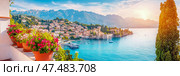 Lake Garda Serenity: A Coastal Italian Village View with Mountains, Boats, and Floral Display. Стоковое AI-изображение, AI-художник Данила Большаков / Фотобанк Лори