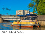 A pleasure catamaran with the inscription "With Us on the River?" and the name "Transbaltika Tour-1", moored at the river bank in Kaliningrad. The boat has sunshades, lifebuoys and is designed for water excursions. In the background is a pedestrian bridge over the Pregolya River with openwork metal decor and passers-by. Behind the bridge are typical Soviet panel houses and green trees. The day is clear, sunny, the water reflects the yellow and blue shades of the city and the sky. (2020 год). Редакционное фото, фотограф Анна Кривицкая / Фотобанк Лори