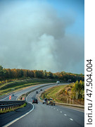 A highway stretching into the distance with a stream of cars moving along it. Behind the road, a dense forest is visible, from behind which a huge column of white-gray smoke rises, indicating a possible forest fire or a large blaze. The contrast between the clear road, greenery and the alarming cloud of smoke creates a sense of impending danger. The sky is partially covered with smoke. Стоковое фото, фотограф Анна Кривицкая / Фотобанк Лори