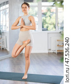 Young girl doing hatha yoga in modern studio, performing balancing asana Vrikshasana with hands clasped in namaste. Стоковое фото, фотограф Яков Филимонов / Фотобанк Лори