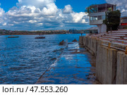 A picturesque view of Sevastopol Bay with a concrete embankment and the steps of an amphitheater facing the sea. Along the shore there is a white building with a terrace, and in the distance you can see ships and cranes of shipyards. The weather is sunny but cloudy, creating a striking contrast between the blue sky, sea and architecture. On the horizon you can see the cityscape of Sevastopol with its historical and industrial sites. (2021 год). Редакционное фото, фотограф Анна Кривицкая / Фотобанк Лори