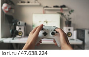 Close-up of hands holding white gaming controller during console gameplay. Blurred screen in background shows immersive action. Modern gaming setup and digital entertainment concept. Стоковое видео, видеограф Енваев Андрей / Фотобанк Лори