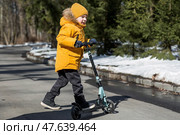 A little boy in bright yellow clothes rides a scooter. A happy child walks outside in early spring. Happy childhood. Стоковое фото, фотограф Photojuli / Фотобанк Лори