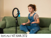 Young female blogger sits on a sofa looking at a phone attached to a ring light. Стоковое фото, фотограф Мария Мороз / Фотобанк Лори
