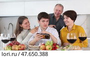 Positive young couple sharing memories with elderly parents sitting at cozy kitchen, browsing photos on smartphone together during festive family meal. Стоковое видео, видеограф Яков Филимонов / Фотобанк Лори