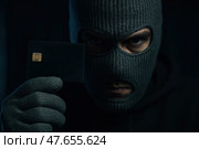 Киберпреступник. Мужчина в балаклаве держит смартфон и кредитную карту в тёмной обстановке. Стоковое AI-изображение, AI-художник Башкирова Анна / Фотобанк Лори