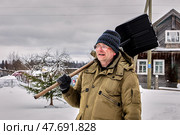 Person wearing blue knit hat and olive green winter jacket carries snow shovel over shoulder while standing in snowy rural area with wooden house and bare trees in background. Стоковое фото, фотограф Владимир Григорьев / Фотобанк Лори