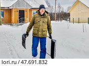 Walking through snowy yard mature man in warm jacket holds large two handed snow pusher in one hand and smaller snow shovel in other. Стоковое фото, фотограф Владимир Григорьев / Фотобанк Лори