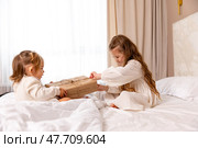 Two sisters wearing white dresses, sitting on a bed with white bedding, pulling big wrapped gift box, fighting, cannot share a present on the holiday morning at home. Стоковое фото, фотограф Ирина Михайличенко / Фотобанк Лори