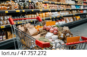 Shopping cart filled with fresh produce and groceries in a supermarket aisle during a busy shopping day. Стоковое AI-изображение, AI-художник Сергей Толмачёв / Фотобанк Лори