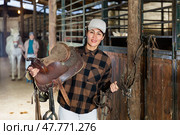 Asian female riding instructor walking in stable with saddle in hands. Стоковое фото, фотограф Яков Филимонов / Фотобанк Лори