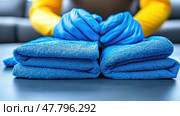 A person in blue gloves is carefully stacking freshly folded blue towels on a table, demonstrating attention to cleanliness and organization. Стоковое AI-изображение, AI-художник Данила Большаков / Фотобанк Лори