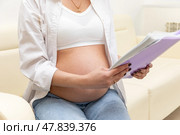 Pregnant woman sitting on sofa in clinic, holding medical record and waiting for doctor's appointment. Happy young expectant mother. Family planning and IVF centers concept. High quality photo. Стоковое фото, фотограф Ирина Михайличенко / Фотобанк Лори