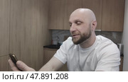 Bearded man talking via video link with his relatives from another city or country. Guy having lively conversation on his smartphone, sitting at home in kitchen. Portrait of young man with phone. Стоковое видео, видеограф Ирина / Фотобанк Лори